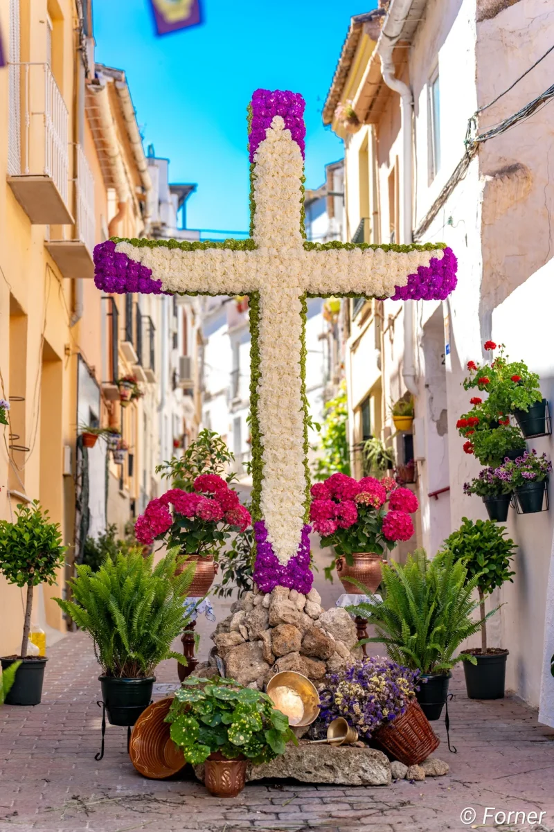 Cruz de flores blancas y moradas entre macetas, en una calle estrecha de casas encaladas