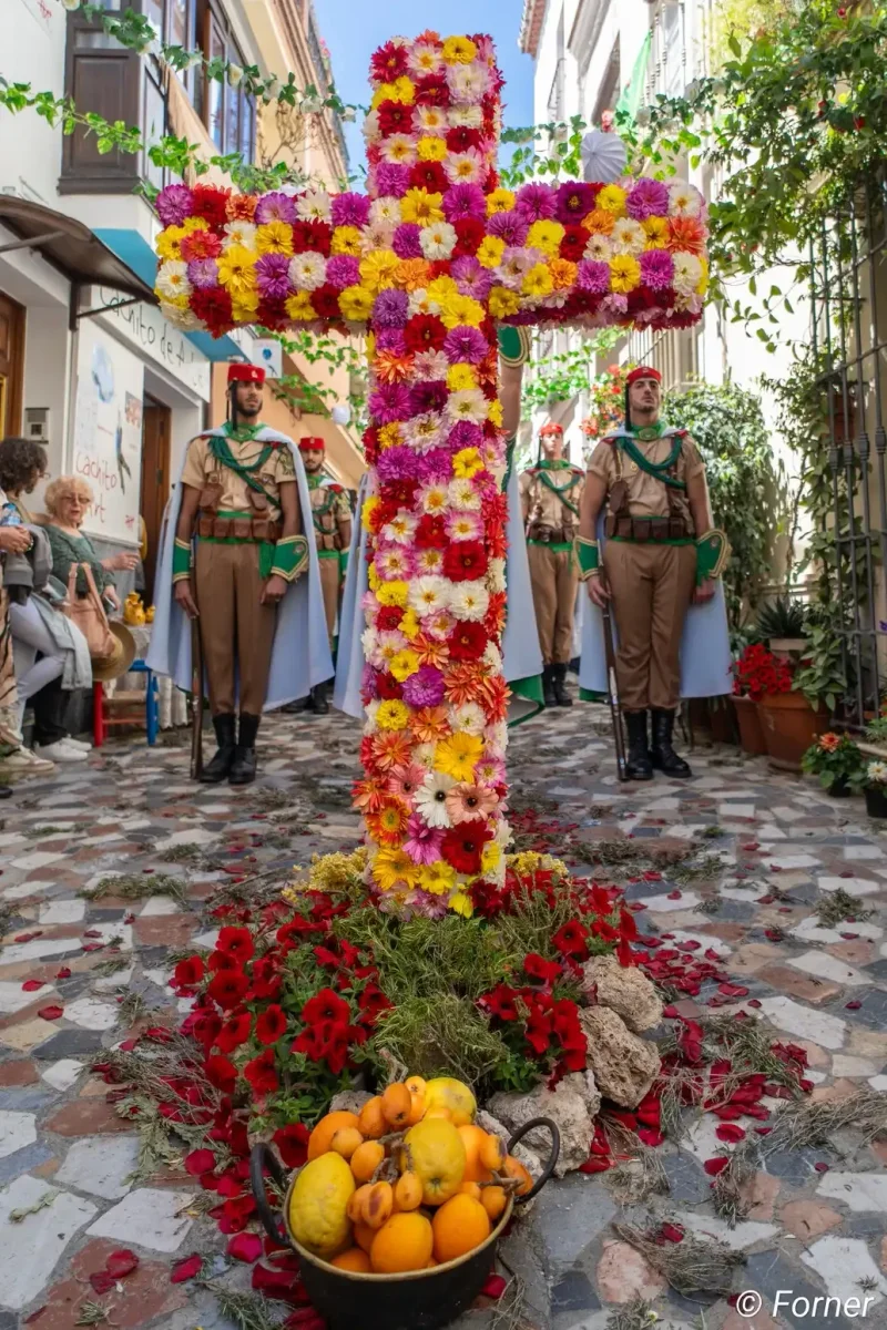 Cruz de flores de colores en una calle empedrada, con frutas a sus pies y guardia de honor