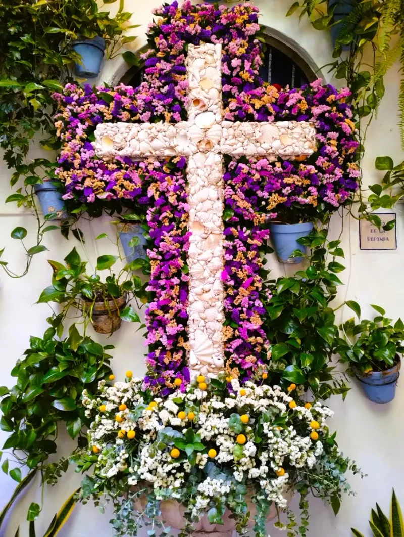 Cruz de conchas sobre un manto de flores moradas y rosas, en una pared con macetas